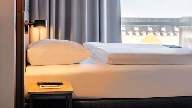 A hotel room with a bed, a nightstand, a lamp, and a view of buildings through the window. - B&B HOTEL Braunschweig-West (Braunschweig)