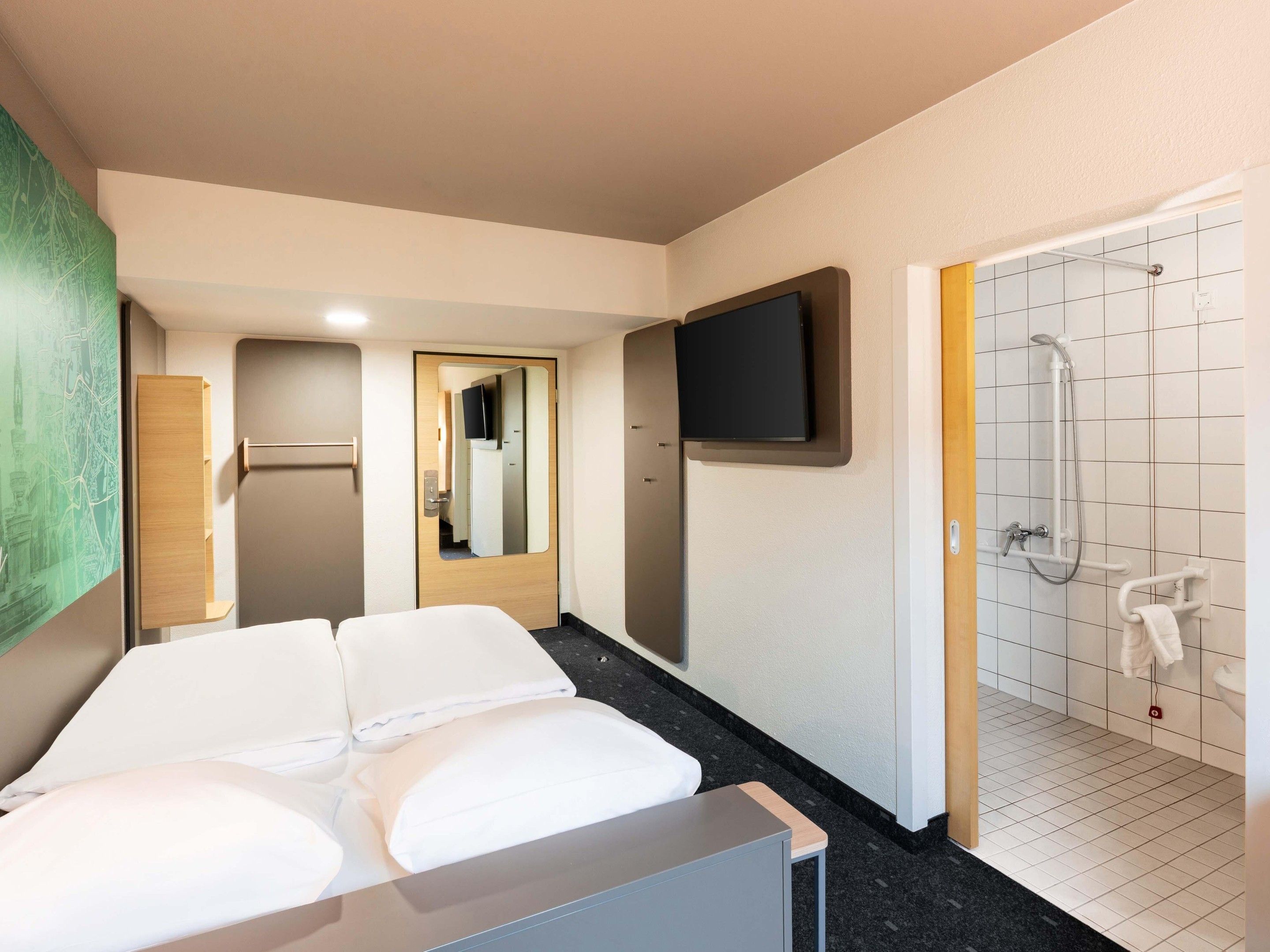A hotel room with a bed, a television mounted on the wall, a shower area with a handheld showerhead, and a wooden cabinet.