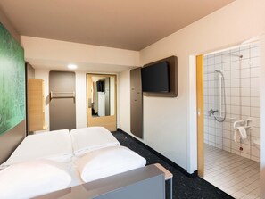 A hotel room with a bed, a television mounted on the wall, a shower area with a handheld showerhead, and a wooden cabinet. - B&B HOTEL Braunschweig-West (Braunschweig)