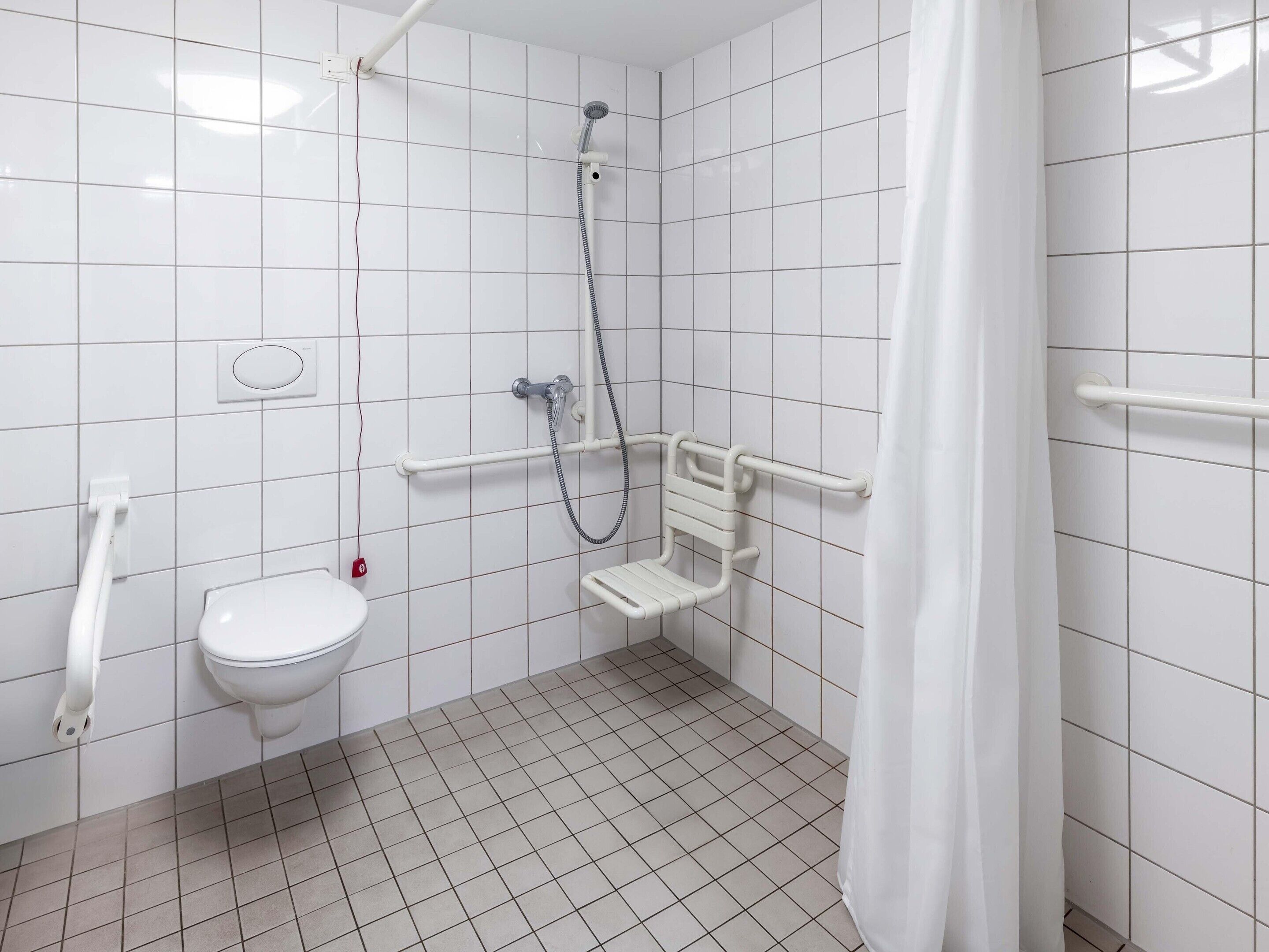 Un baño con azulejos blancos, un inodoro, una ducha con barra de apoyo y un cortinón blanco.