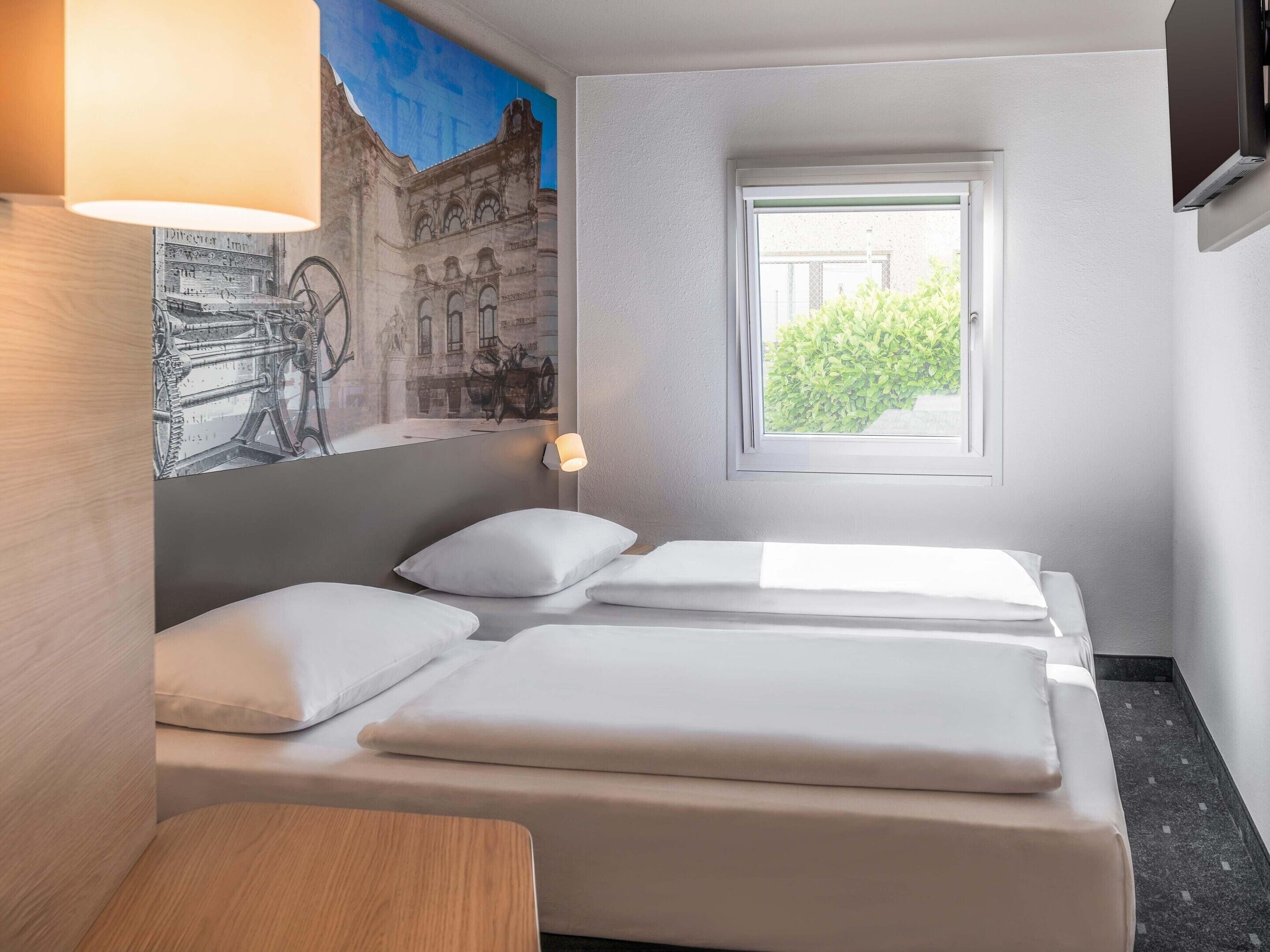 A hotel room with two beds, a small table, a wall-mounted TV, and a window with a view of greenery.