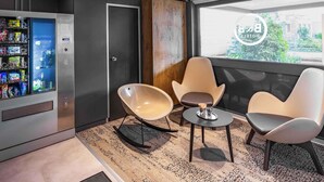 Una habitación de hotel moderna con una máquina expendedora, dos mecedoras, una mesita y una ventana con vistas a edificios y árboles.
