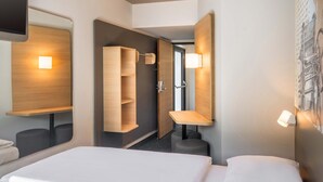 Una habitación de hotel con cama, espejo, estante de madera, lámpara fijada a la pared y puerta que da a un baño.
