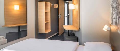 A hotel room with a bed, a mirror, a wooden shelf, a wall-mounted lamp, and a door leading to a bathroom.