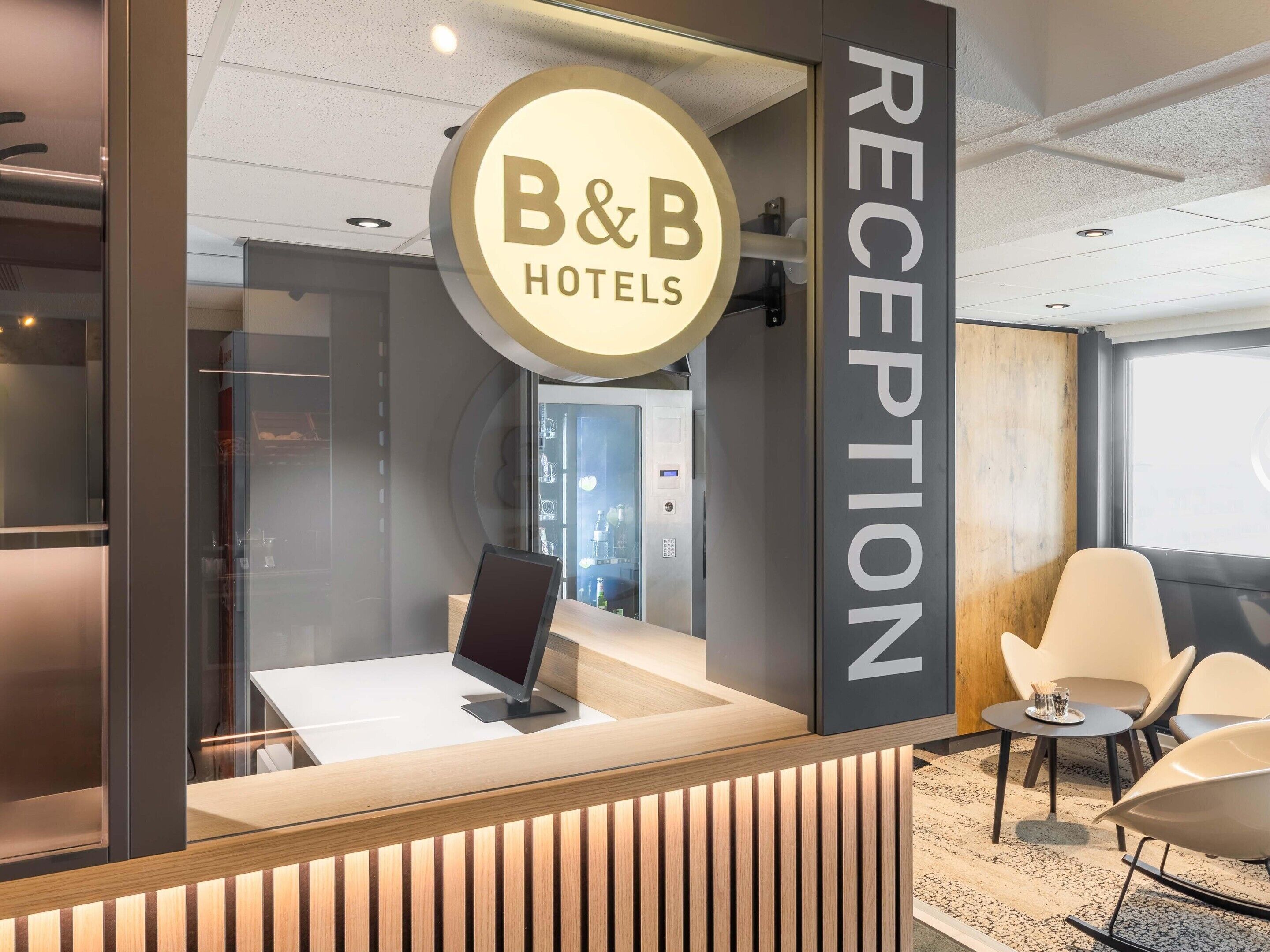 Uma área de recepção com uma placa da B&B Hotels, um balcão de atendimento e assentos modernos.