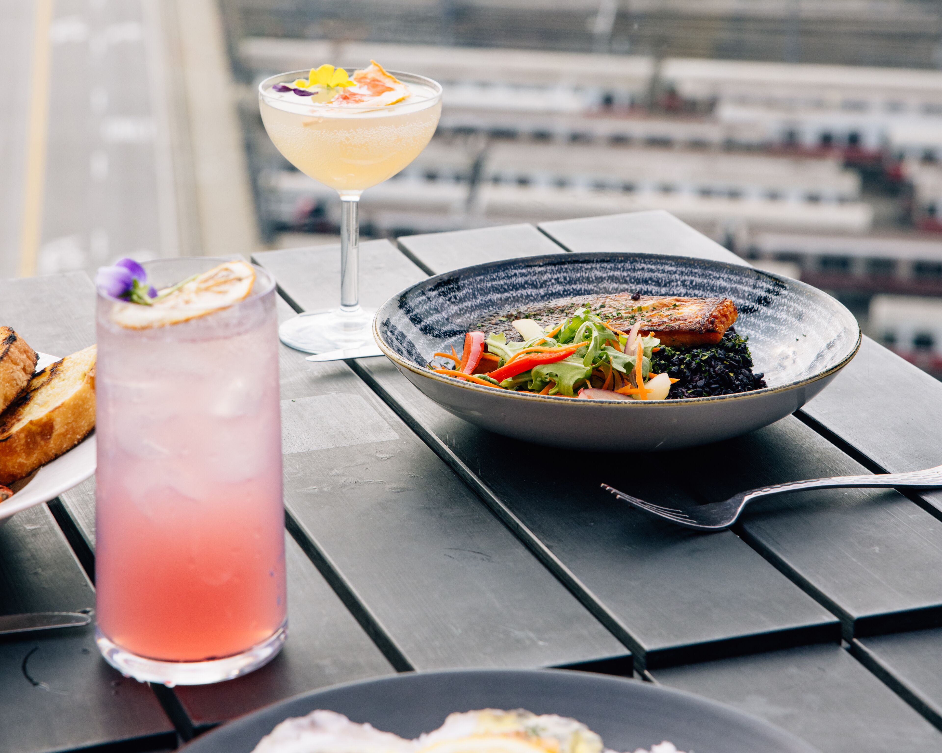 A meal with fish and vegetables on a plate, a glass of pink drink, and a slice of bread on a table.