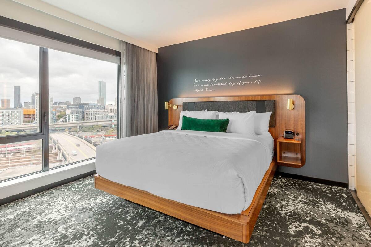 Habitación de hotel con un ventanal grande, cama con ropa de cama blanca, almohada verde y cabecero de madera, además de una cita motivacional en la pared.