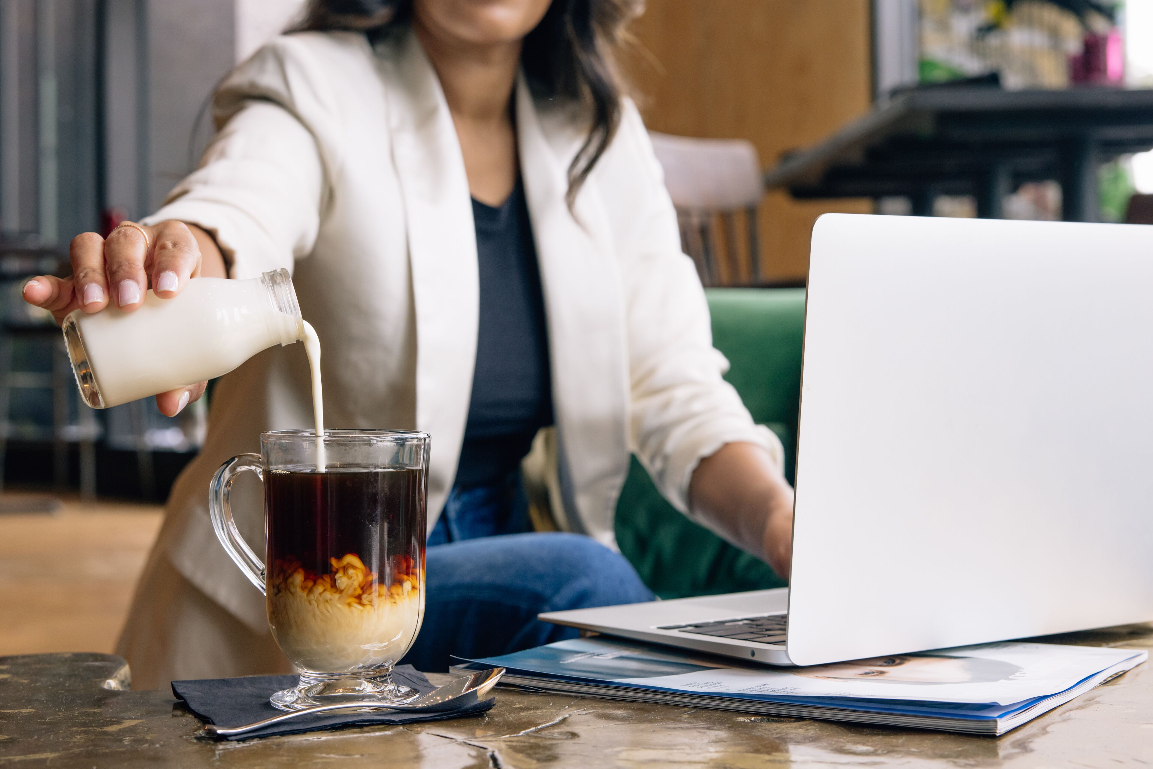 A person pouring milk into a cup of coffee on a table with a laptop and documents.