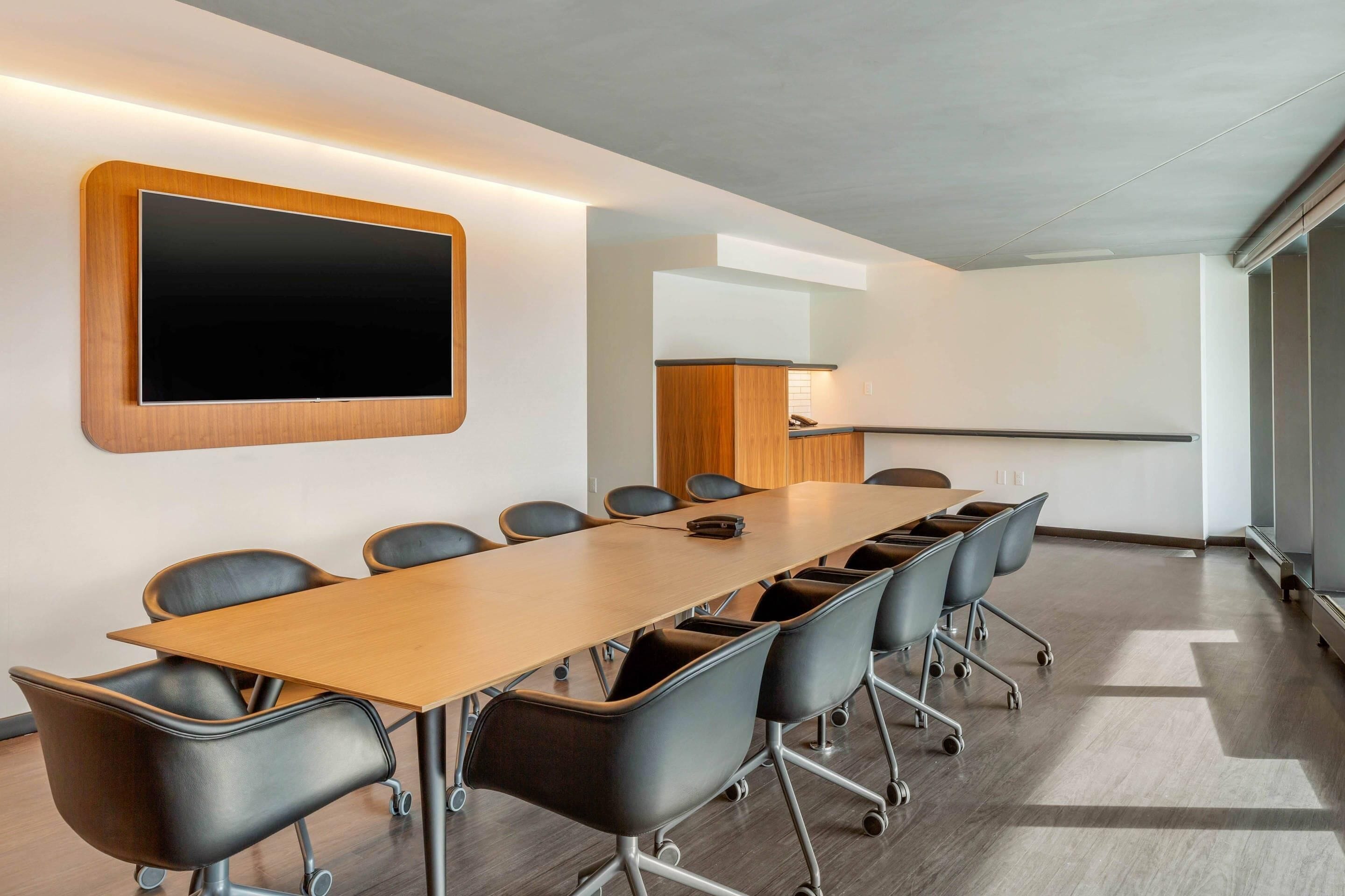 Un salón de conferencias con una larga mesa, sillas negras, un televisor montado y una pared de madera decorativa.