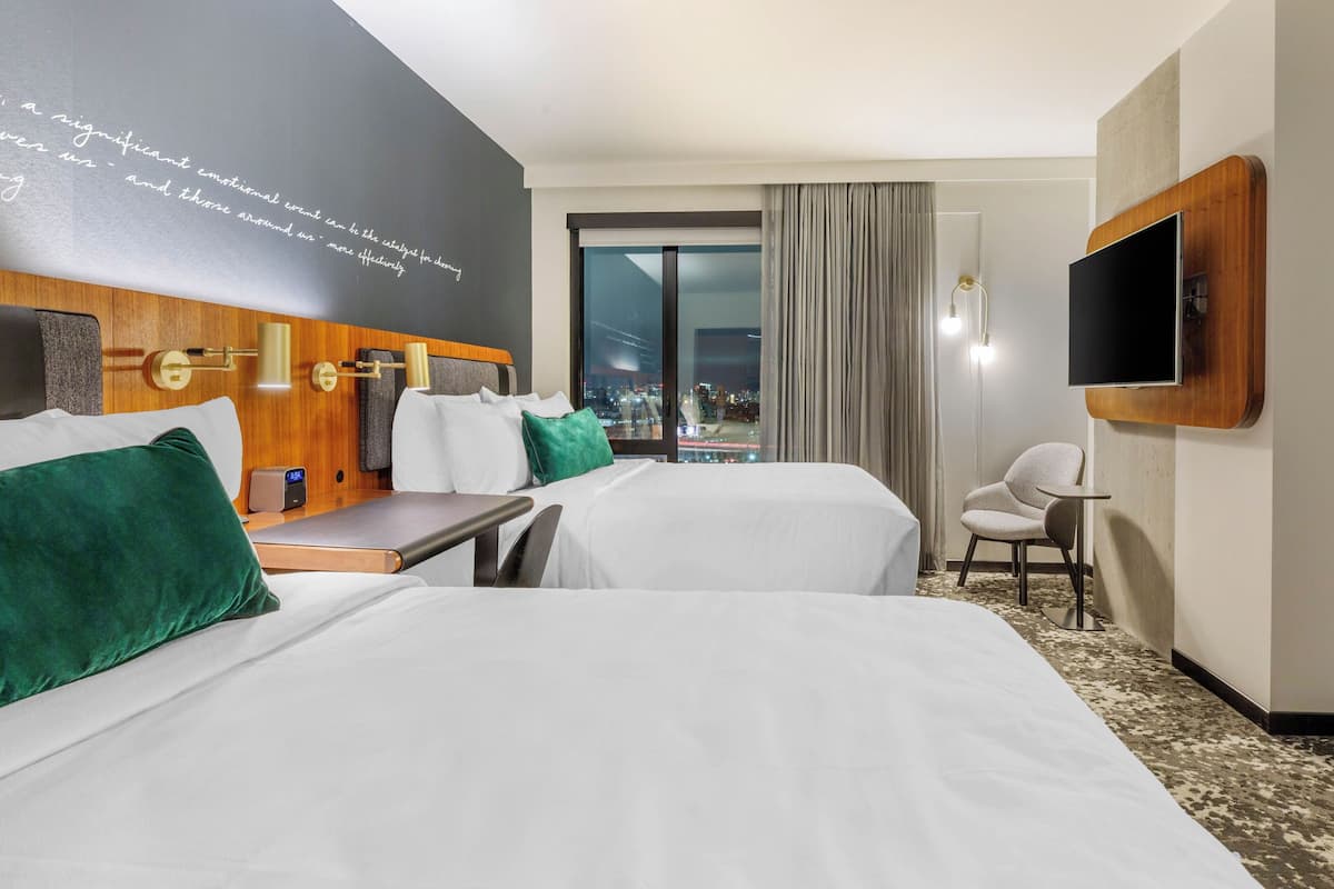 Habitación de hotel con dos camas, un escritorio, una silla, un televisor y un ventanal con vistas a la ciudad.