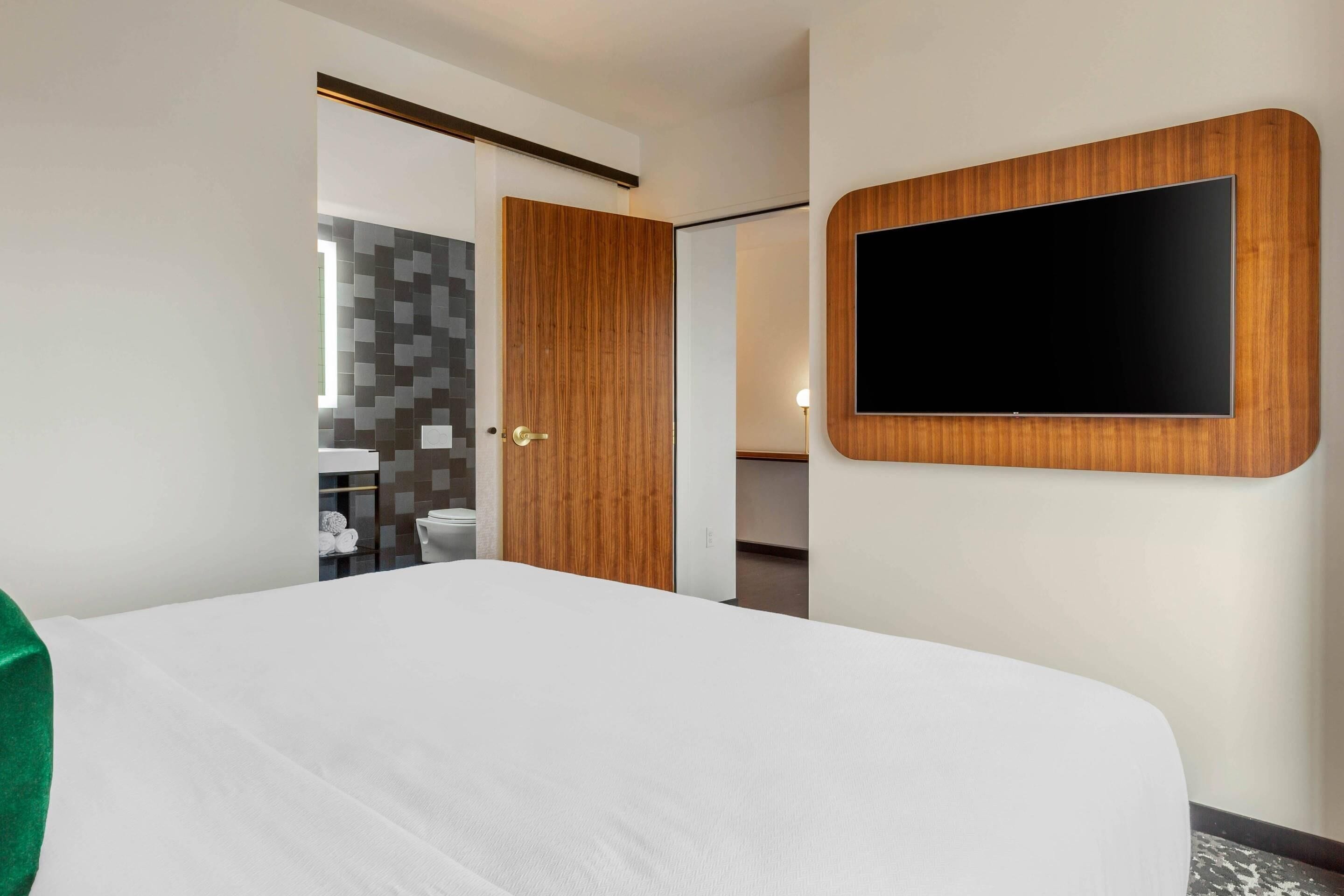 A hotel room with a bed, a TV mounted on the wall, a wooden door, and a bathroom visible through an open door.