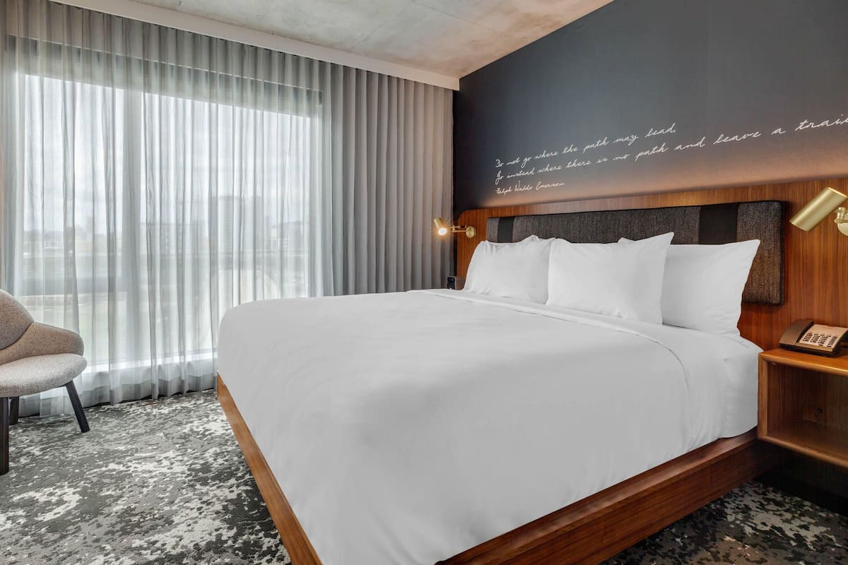 Habitación de hotel con una cama grande, una silla, una mesita de noche con un teléfono y una pared con una cita.