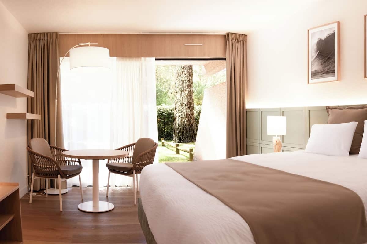 A hotel room with a large bed, a desk with chairs, a view of the outdoors, and framed pictures on the wall.