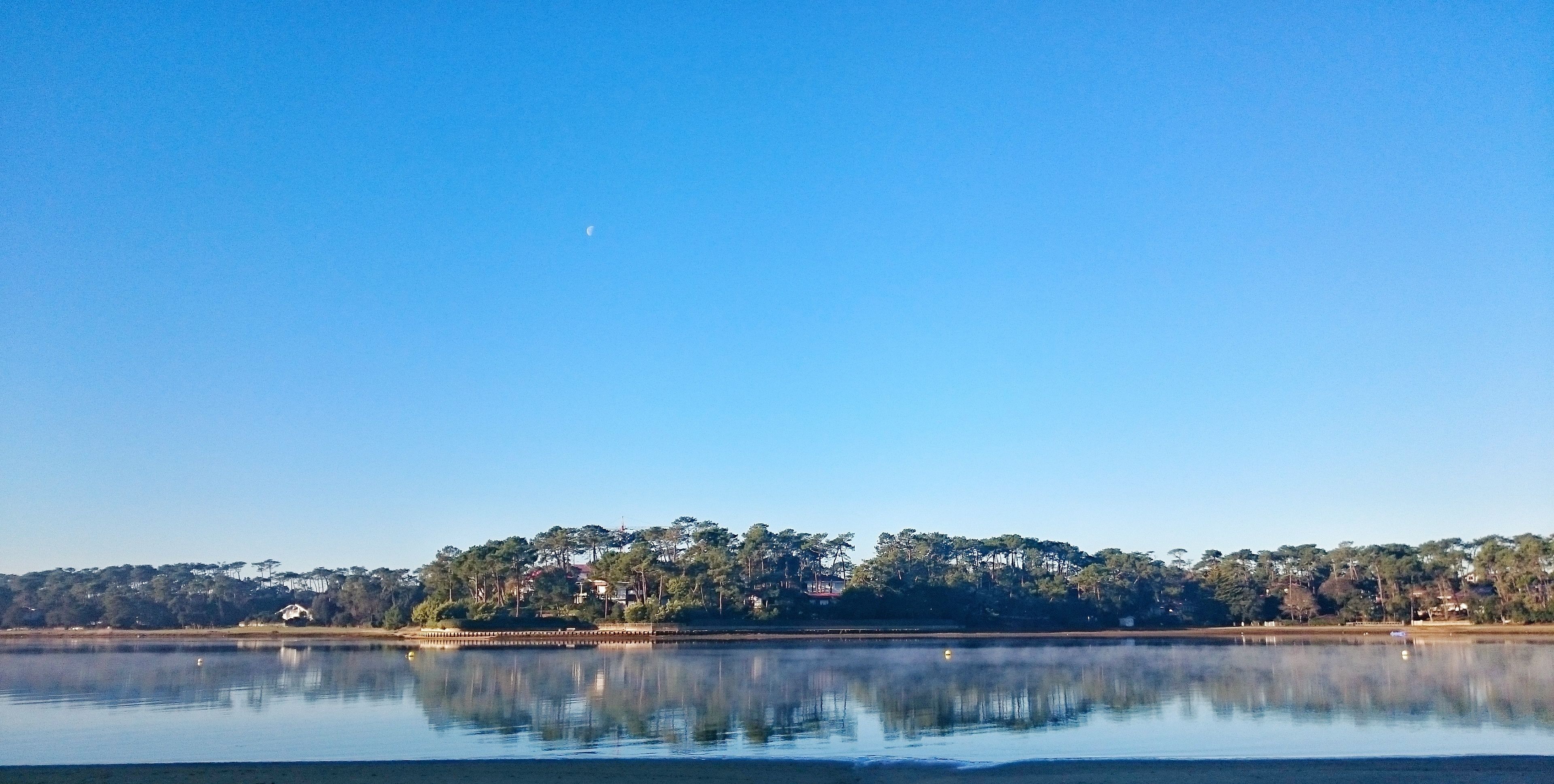 Ein ruhiger See mit klarem Himmel, einem sichelförmigen Mond und einer bewaldeten Uferlinie mit Gebäuden.