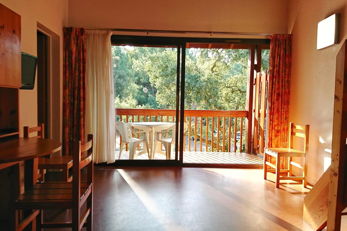 A room with a wooden floor, a dining table with chairs, a staircase, and a balcony with a view of trees.