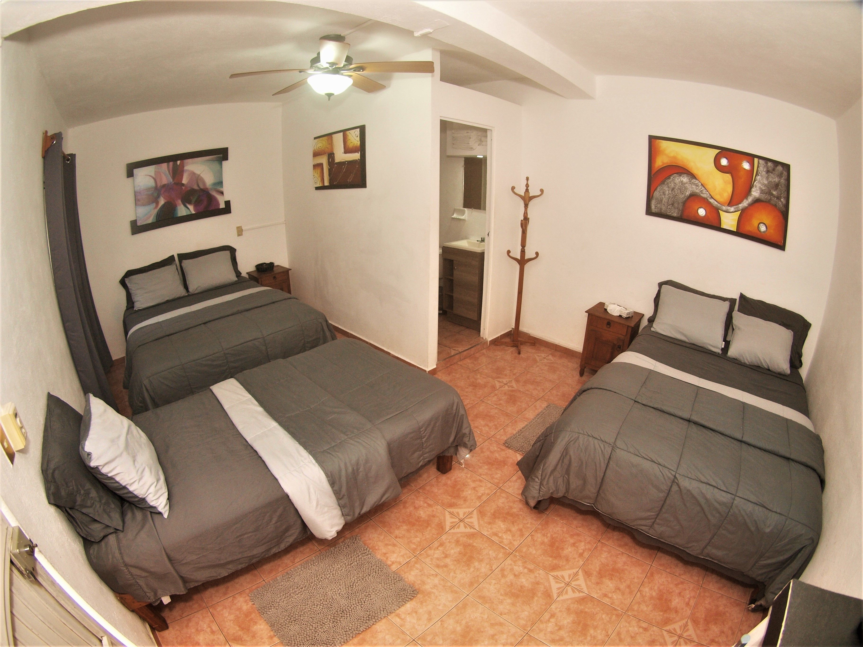 Una habitación con dos camas, ventilador de techo, obras de arte en las paredes y un baño visible a través de una puerta abierta.