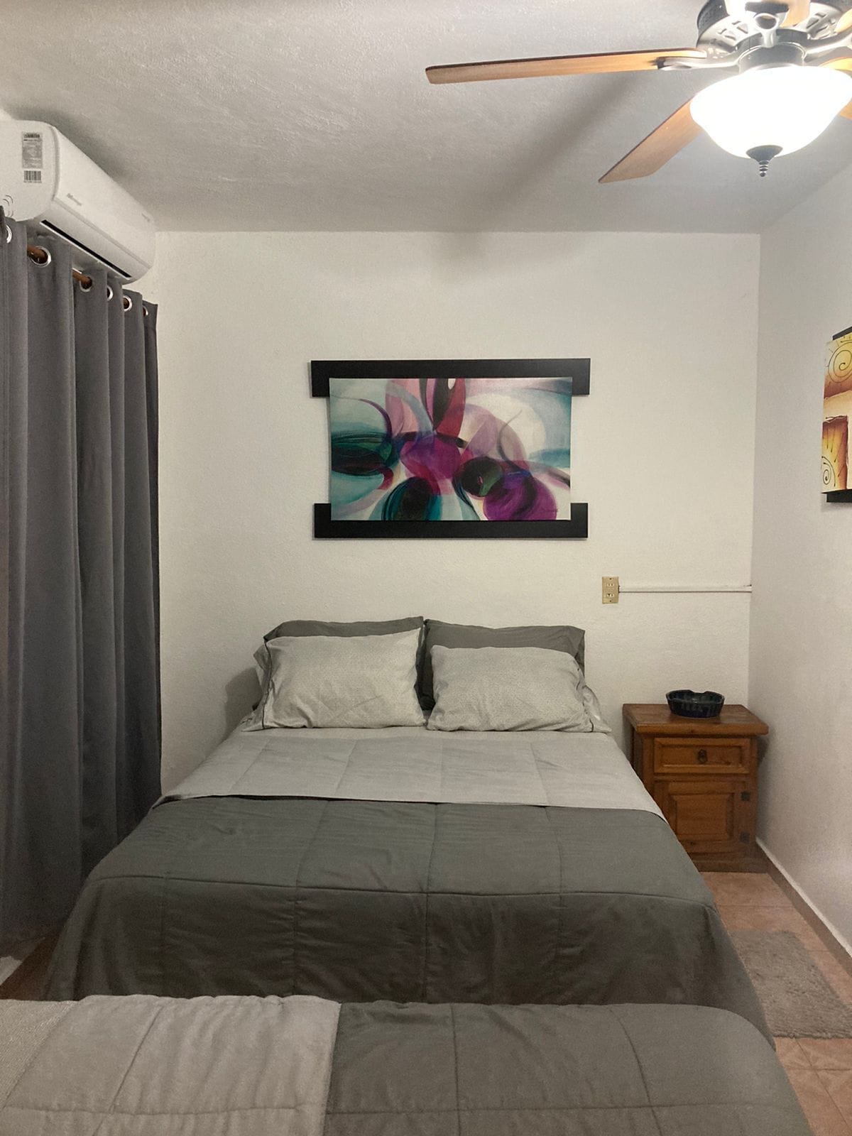A bedroom with a bed, a ceiling fan, a wall-mounted air conditioner, a nightstand with a bowl, and a painting on the wall.