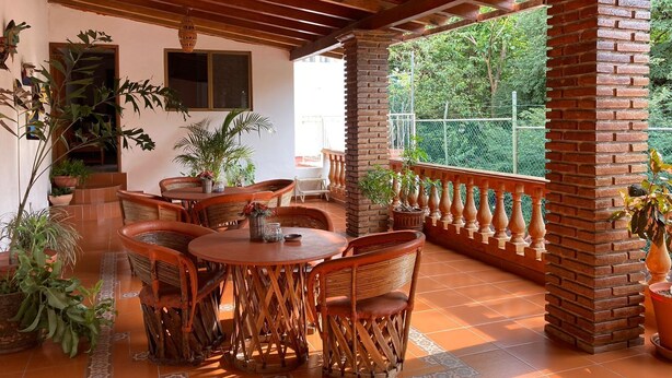 A covered patio with a round wooden table and wicker chairs, surrounded by potted plants and a brick column. - Hostal Casa Blanca (Comala)