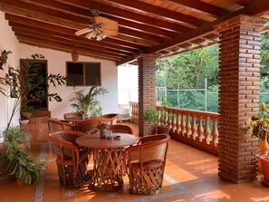 Un patio cubierto con una mesa redonda de madera y sillas de mimbre, rodeado de macetas con plantas y un pilar de ladrillo.