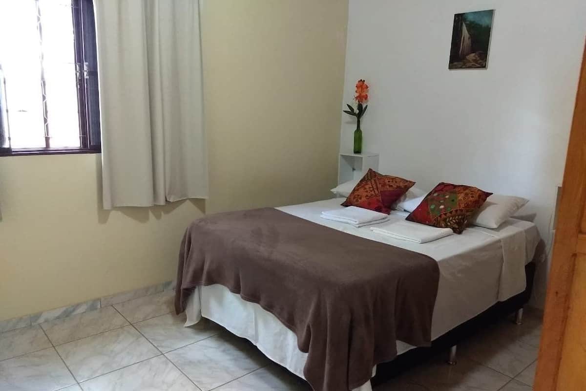 A bedroom with a bed, pillows, a vase with flowers, a painting on the wall, and a window with curtains.