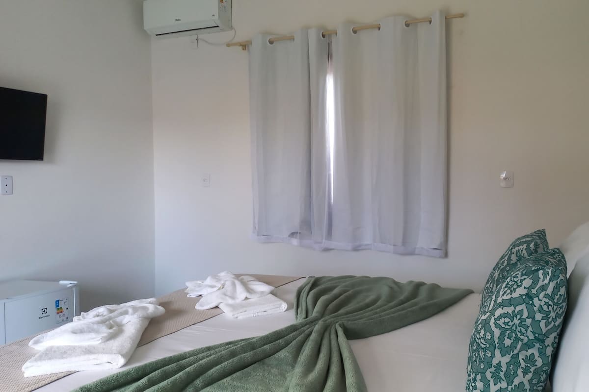 A hotel room with a bed, a green blanket, white towels, a patterned pillow, a TV, and an air conditioning unit.