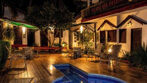 An outdoor pool area with seating and lounge chairs, surrounded by greenery and a building with a sign. - Pousada Quinta Dos Quintais (Chapada dos Guimarães)