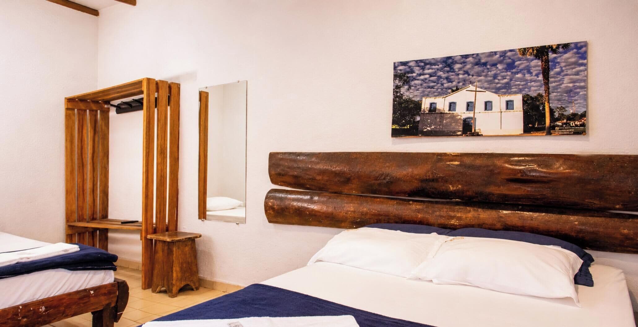 Una habitación de hotel con cabecera de madera, una cama con sábanas blancas, un banco de madera, un espejo y un cuadro de un edificio.