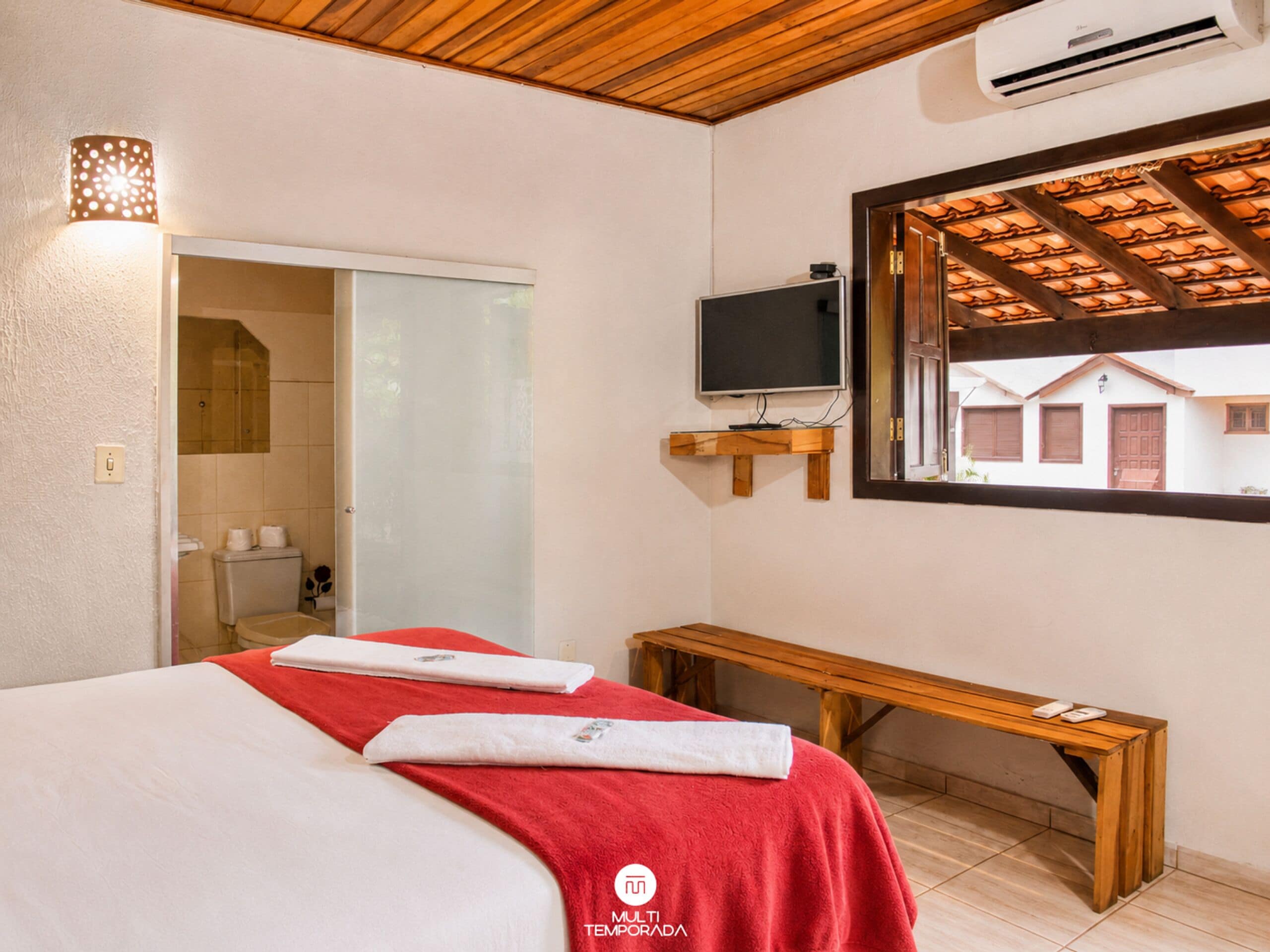 Habitación de hotel con cama, televisor, banco y ventana con vista a edificios.