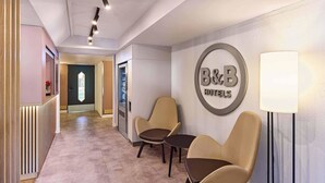 Un espace d’accueil d’hôtel avec une enseigne indiquant « chambre d’hôtes », deux fauteuils en cuir, une lampe sur pied et un comptoir de réception.