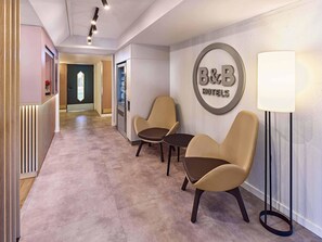 Zona de recepción de un hotel con un cartel de B&B, dos sillones de cuero, una lámpara de pie y un mostrador de recepción.