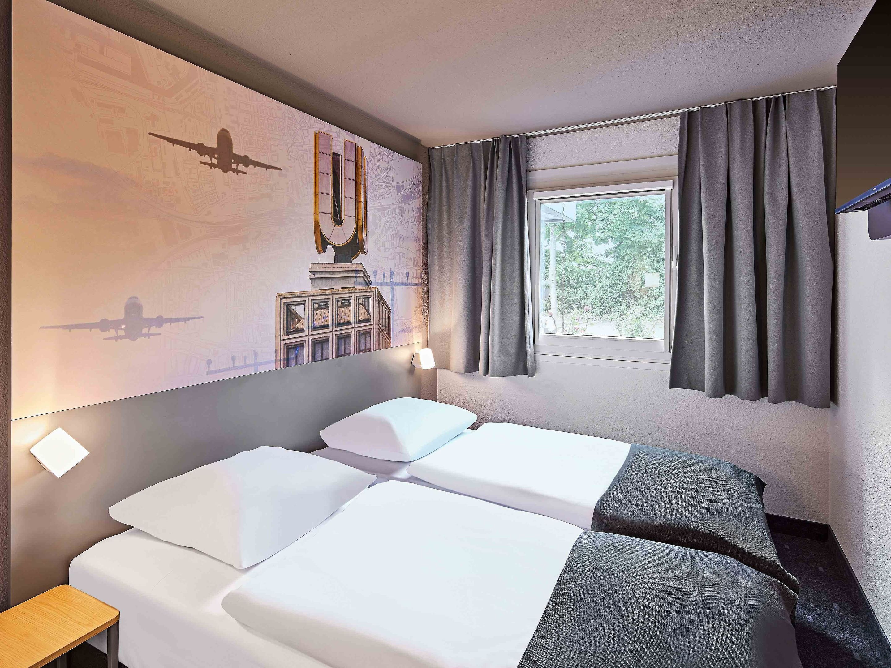A hotel room with two beds, a window with curtains, a wall mural, and a small table.