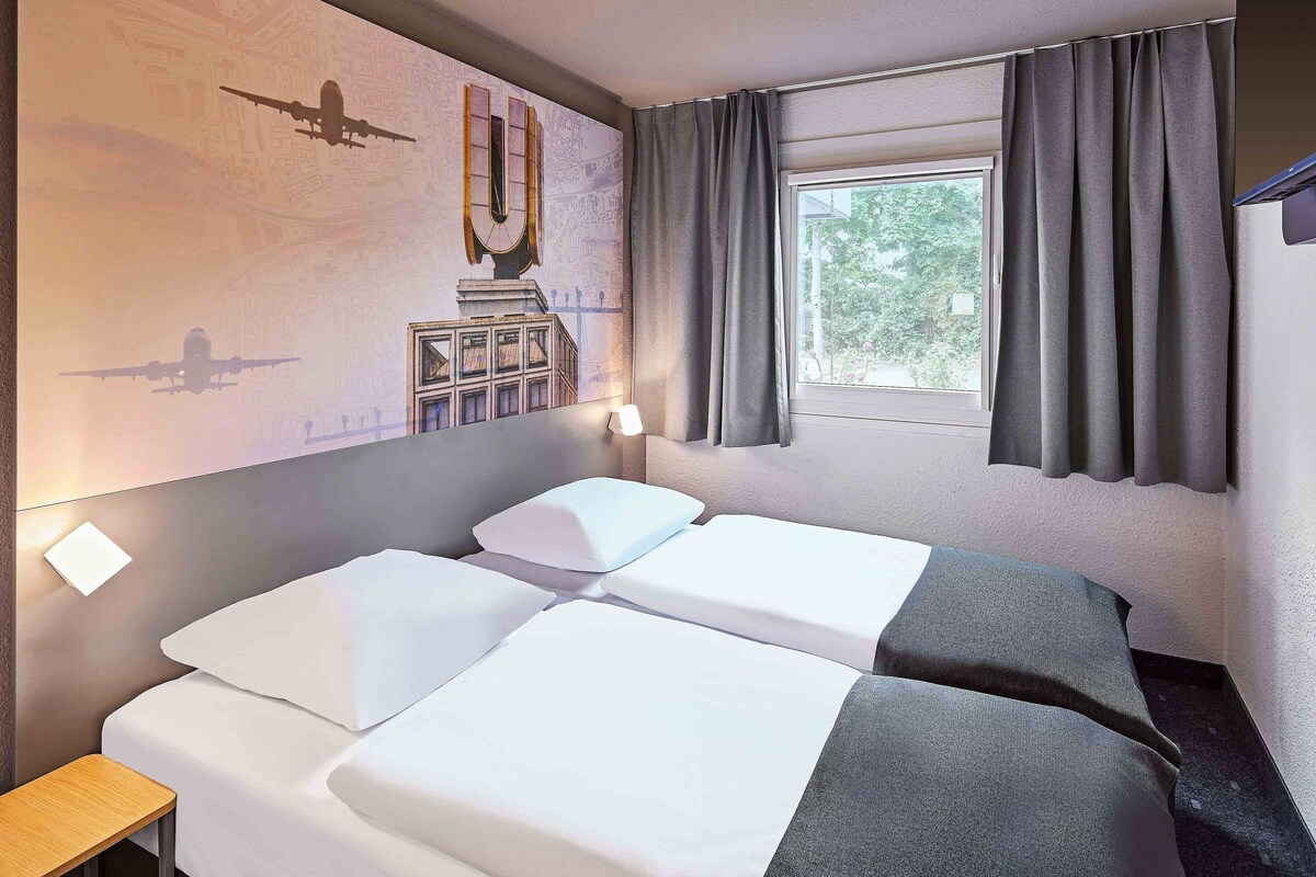 Habitación de hotel con dos camas, una ventana con cortinas, un mural en la pared y una mesita.