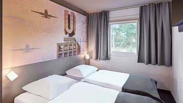 A hotel room with two beds, a window with curtains, a wall mural, and a small table.