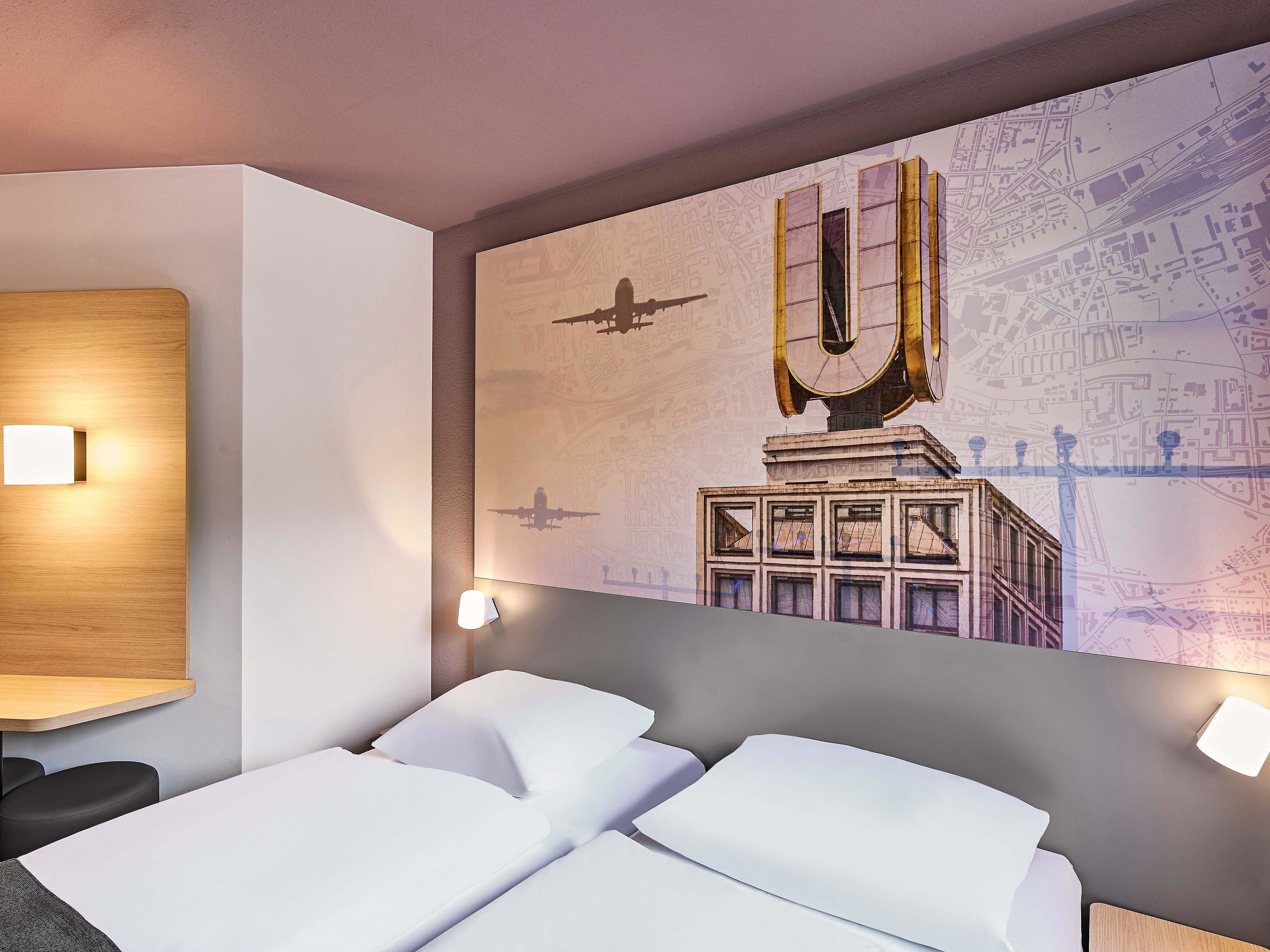 A hotel room with a large bed, two bedside tables, and a wall mural depicting a cityscape with a large 'U' structure.