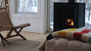 A cozy room with a fireplace, a wicker chair, and a view of trees through the window. - Downtown! Huge 3,000 sq. ft Historic CVX Cottage, Sleeps 16! New Central A/C!!! (Charlevoix)