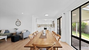 Een ruime eethoek met een houten tafel en stoelen, een keuken op de achtergrond en grote ramen met uitzicht op de omgeving.