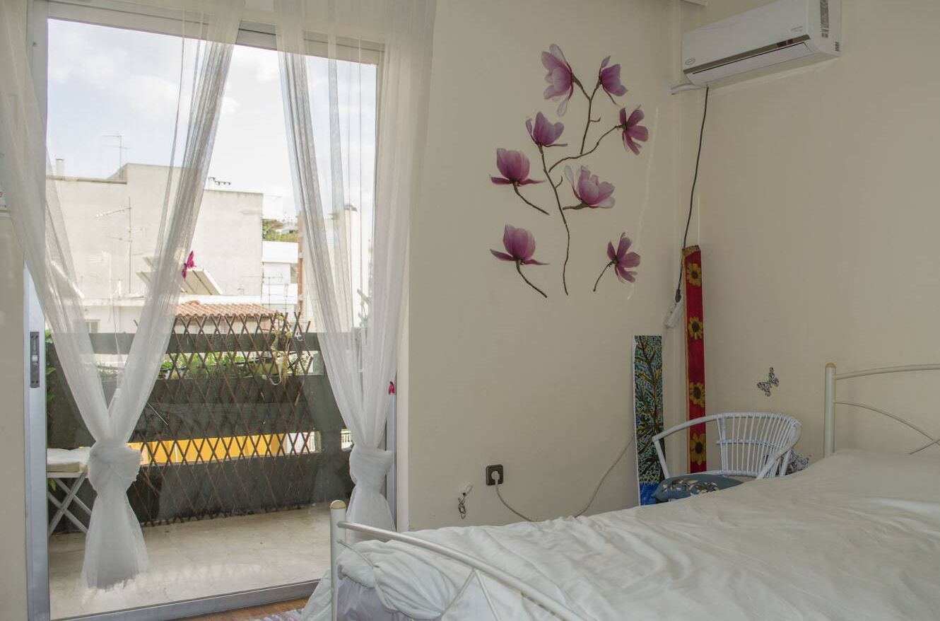 A bedroom with a bed, a window with sheer curtains, an air conditioning unit, and a wall-mounted flower decoration.