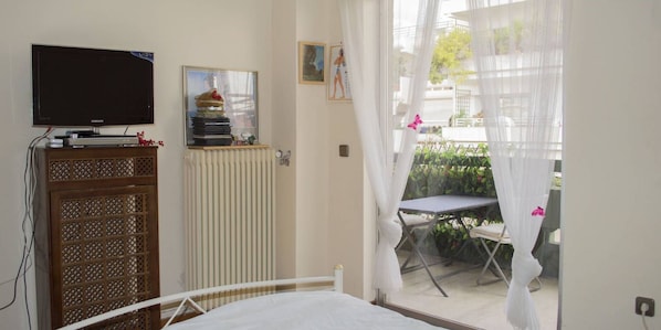 Ein Schlafzimmer mit Bett, einem Fernseher auf einer Kommode, einer Heizung, einem Fenster mit leichtem Vorhang und Blick auf einen Balkon mit Tisch und Stühlen.