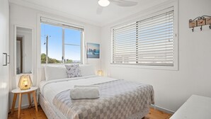 Une chambre à coucher avec un grand lit, deux fenêtres équipées de stores, une table de chevet avec une lampe et une œuvre d’art accrochée au mur.