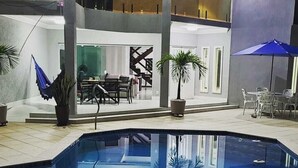 Een modern zwembad met een terras, buitenmeubels en een blauwe parasol, met een tweelaags gebouw op de achtergrond.