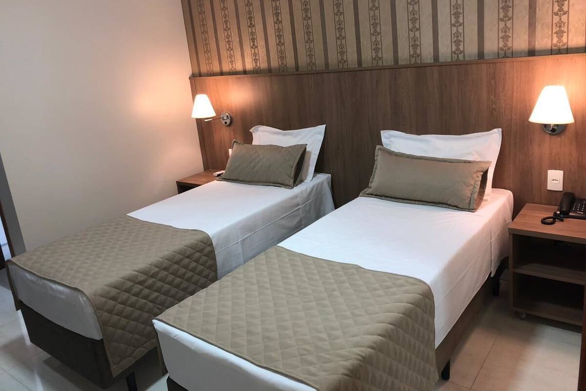 A hotel room with two single beds, each with a quilted bedspread and pillows. There are bedside tables with lamps and a phone on one side. The room has a wooden headboard and a patterned wallpaper on the wall.