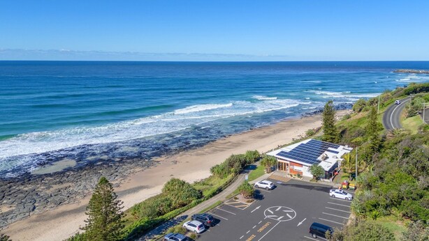 A coastal road with a parking lot, a building with solar panels, and a beach with rocky shoreline. - Surfside - Shelly Beach Holiday Home (East Ballina)