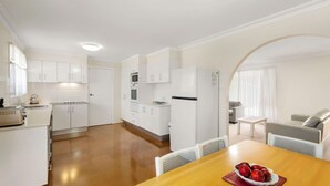 A modern kitchen with white cabinets, a dining area with a wooden table, and a living space with a sofa and a TV.