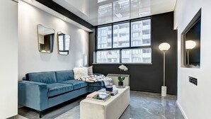 Una sala de estar moderna con un sofá azul, un reposapiés blanco y un ventanal con vistas a un edificio.