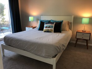 Cama bem arrumada com cabeceira branca, duas mesas de cabeceira com abajures e vista para o exterior através de uma janela com cortinas.
