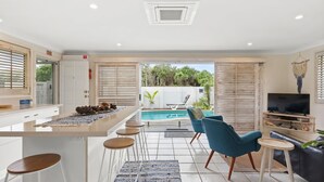 Una cocina moderna con una isla central, taburetes de bar, un área de piscina con una tumbona y un televisor sobre un soporte de madera.