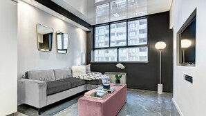 A modern living room with a grey sofa, a pink ottoman, and a large window with a view of buildings.