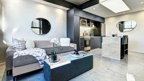 Een moderne woonkamer met een grijze bank, een salontafel, en een keukenhoek met een marmeren aanrecht en roestvrijstalen apparatuur.