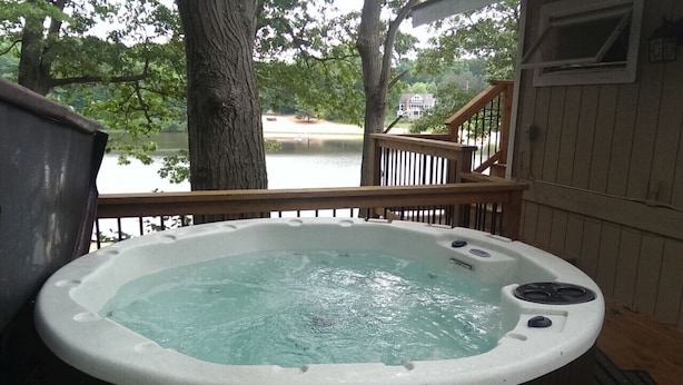A hot tub on a wooden deck with a view of a lake and a house in the background. - Cute Beachfront Guest Unit on Lake (Saugatuck)