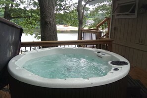 Un bain à remous sur une terrasse en bois, avec vue sur un lac et une maison en arrière-plan.